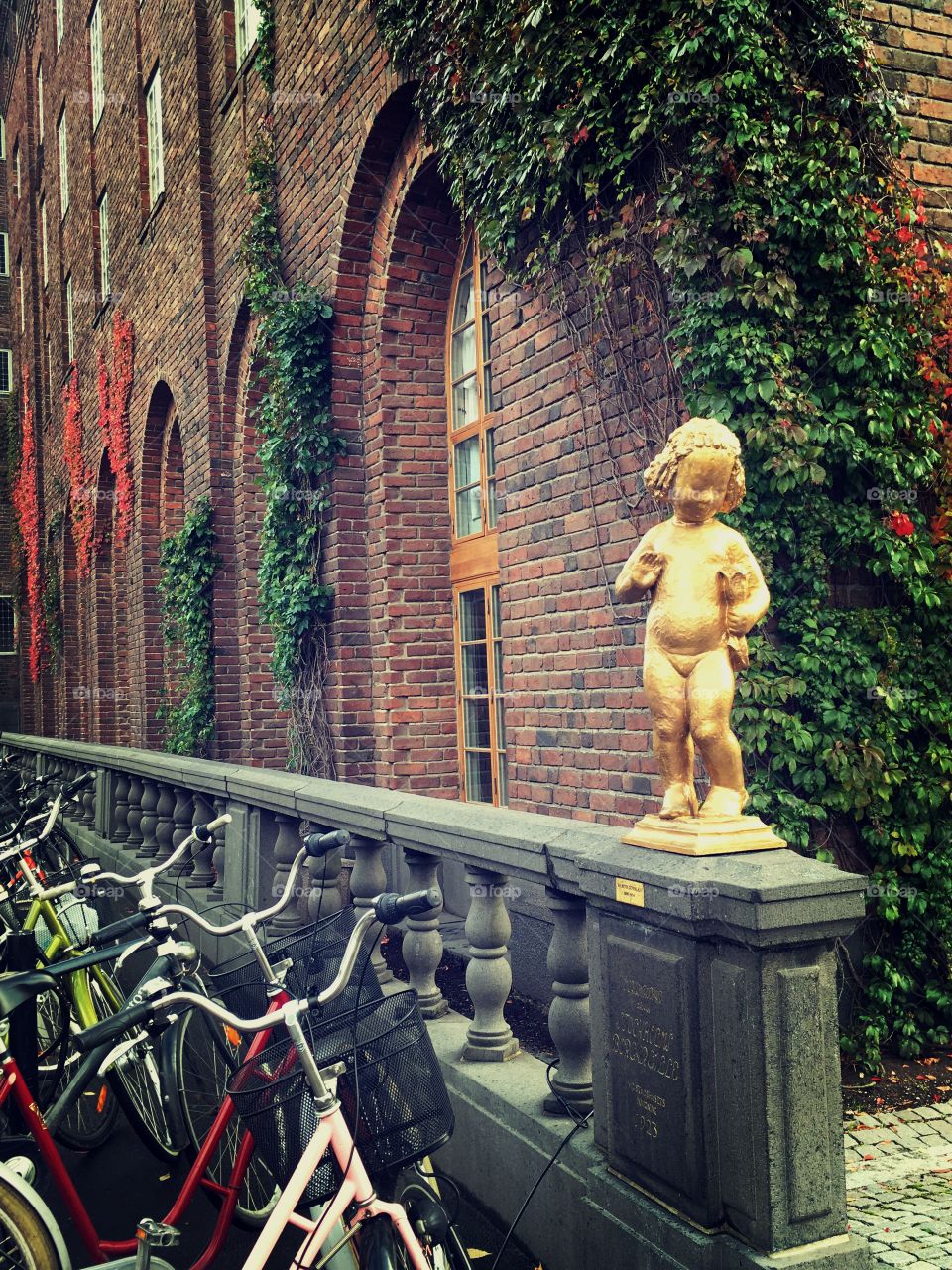 A brick building in Stockholm covered in vines and leaves with a row of bicycles leaning against it. And a golden statue of a baby 