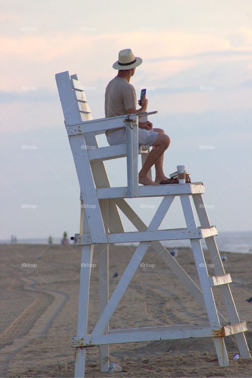 A man in a hat, sitting in a lifeguard chair along the beach at dawn. 