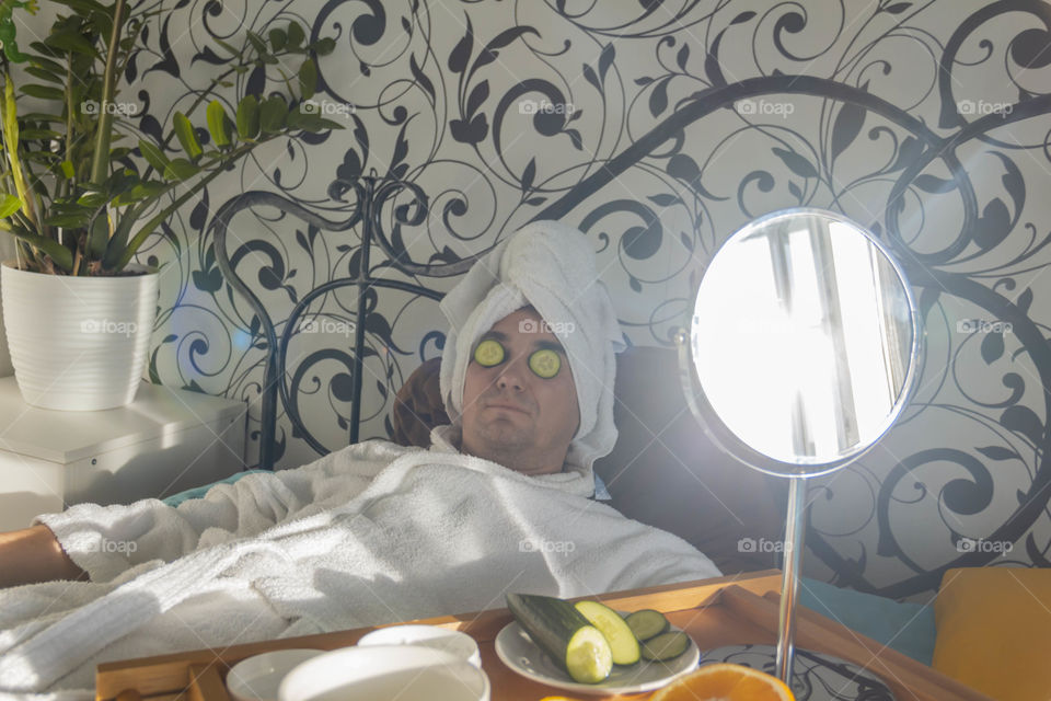 A man and home spa care at home takes care of himself and smears his face with cream, does a manicure at home on a blue background and with oranges and rests.