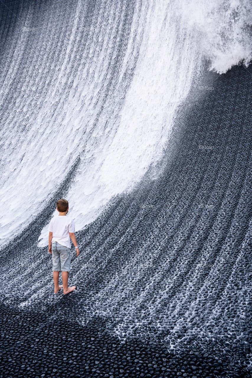 A boy in denim shorts and a white T-shirt looks at an artificial waterfall. Creative fountain in the form of a waterfall. Minimalistic photo in gray tones. Conceptual water installation. Expo 2020 Dubai