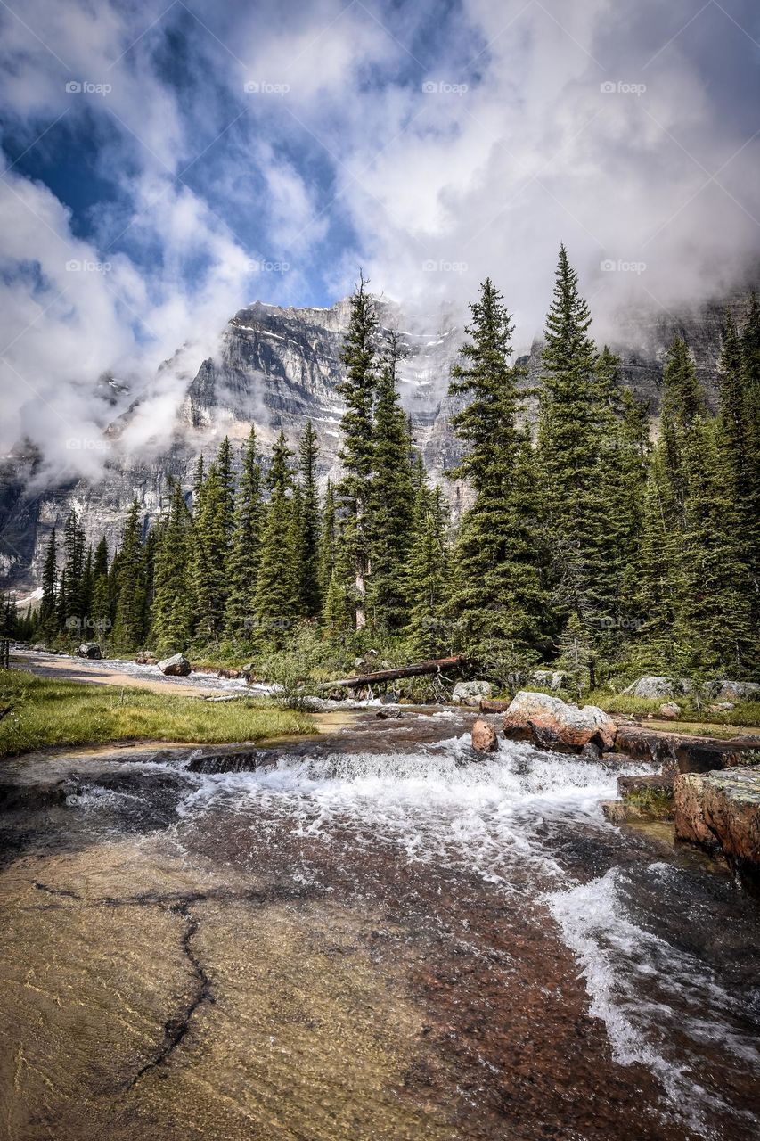 Water rushes down a shallow slope as a dramatic scene of conifers, mountains and low smoky clouds unfold