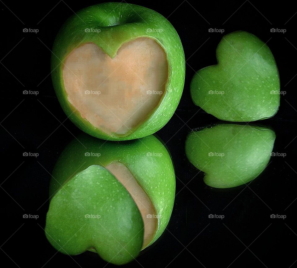 Green color.  On a black mirror background lies a green apple from the surface of which a part in the form of a heart is cut off.  Nearby is a green heart made of an apple.  In the reflection is a green apple with a piece of a green apple heart