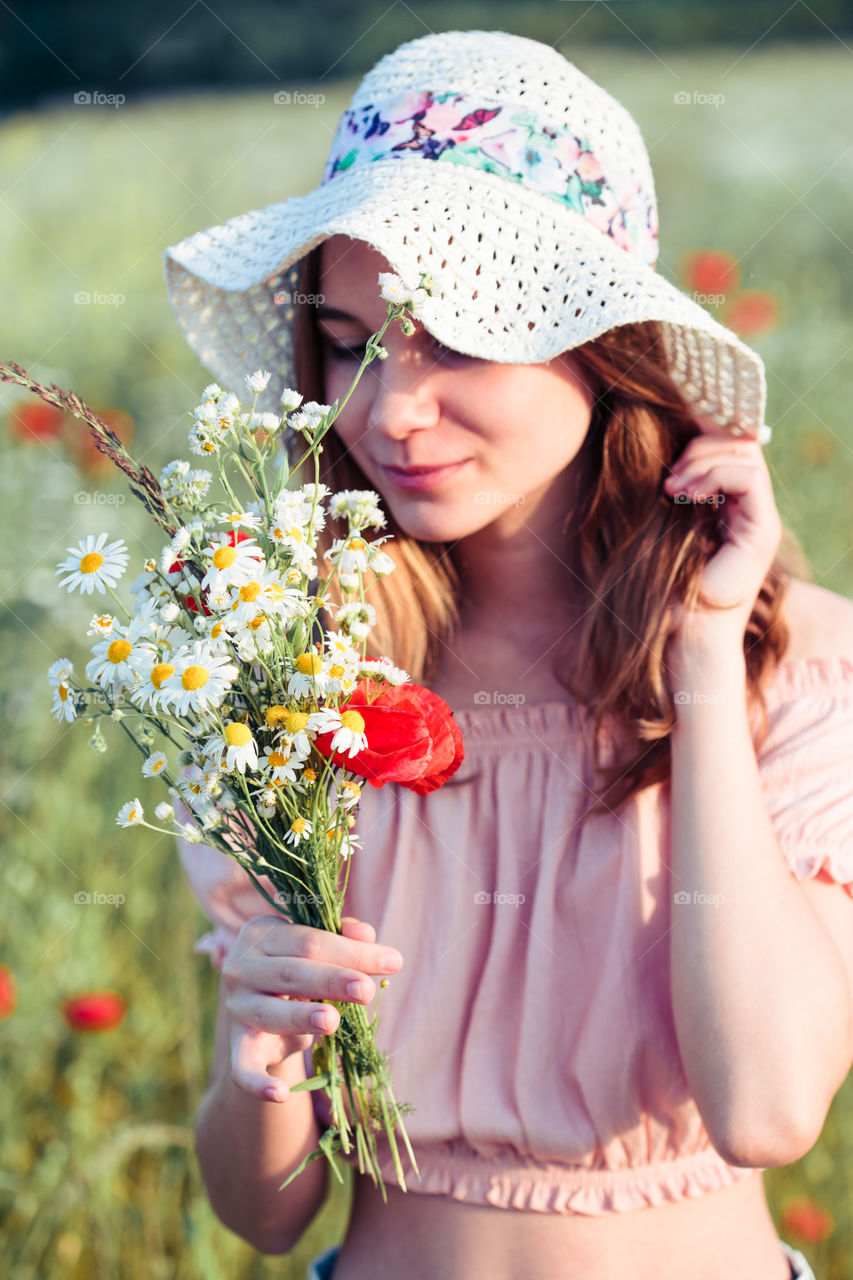 Beautieful young girl in the field of wild flowers. Teenage girl picking the spring flowers in the meadow, holding bouquet of flowers. She wearing hat and summer clothes. Spending time close to nature