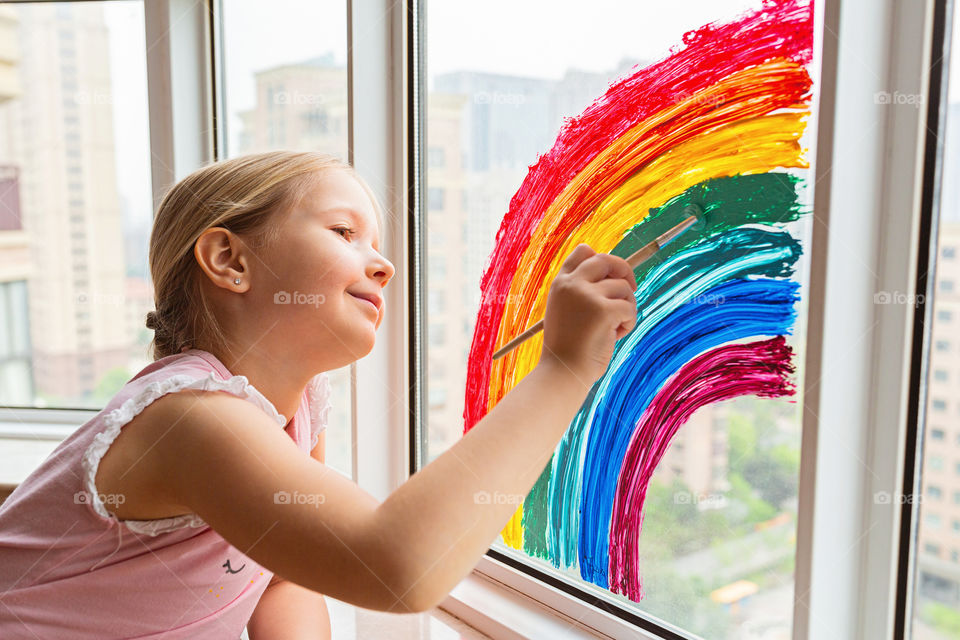 Happy little Caucasian girl with blonde hair painting rainbow during COVID-19 coronavirus quarantine and self isolation 