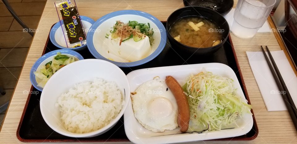 A healthy brunch in Japan, which consists of a bowl of white rice, fried egg, sausage, salad, cold tofu, seaweed and miso soup.