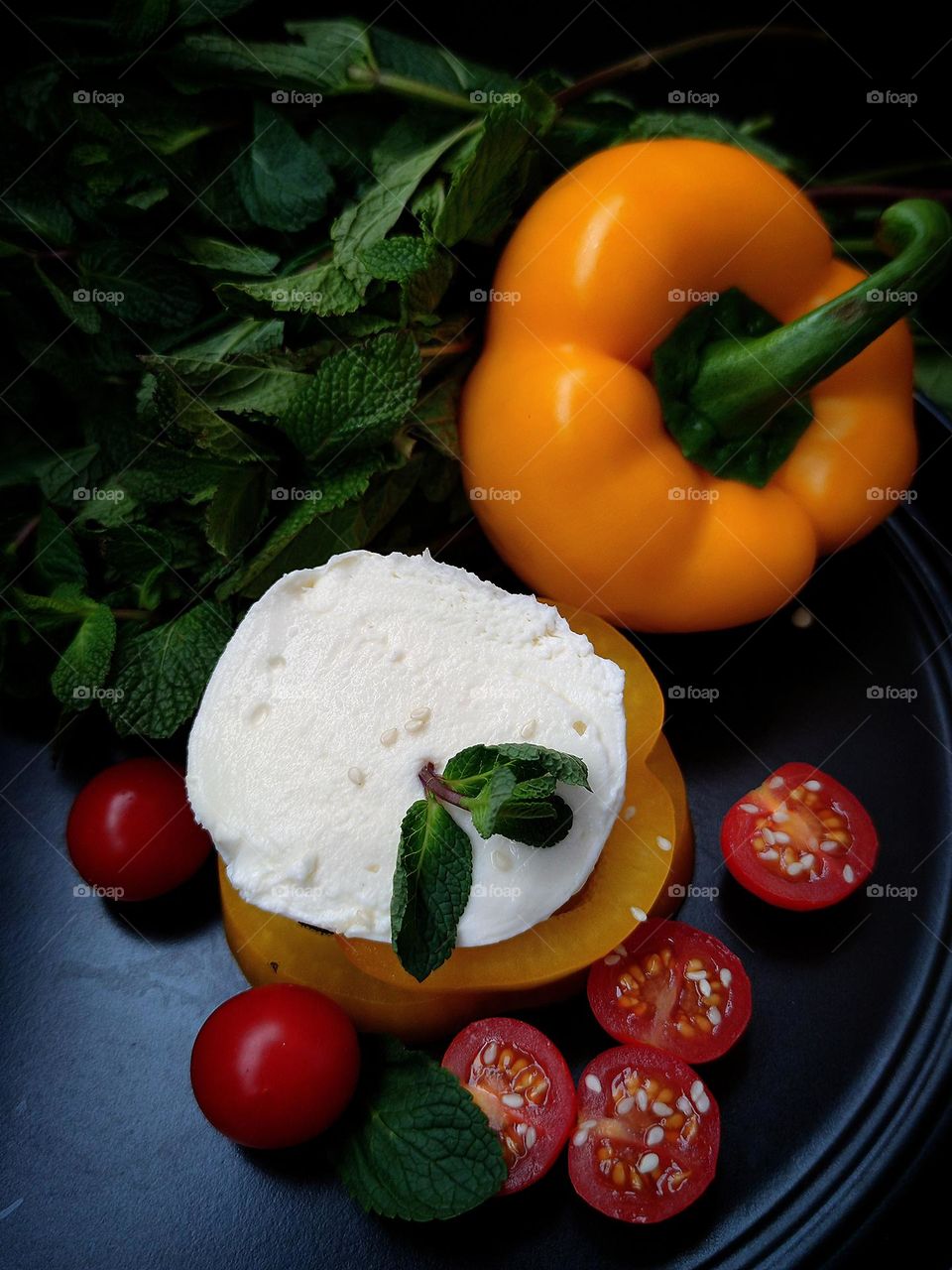 Still life: white burrato cheese lies on mugs of yellow bell pepper.  Nearby are cut red tomatoes and red tomatoes, yellow bell pepper, which closes a bunch of green mint.  Cheese and tomatoes sprinkled with sesame seeds.  Black background