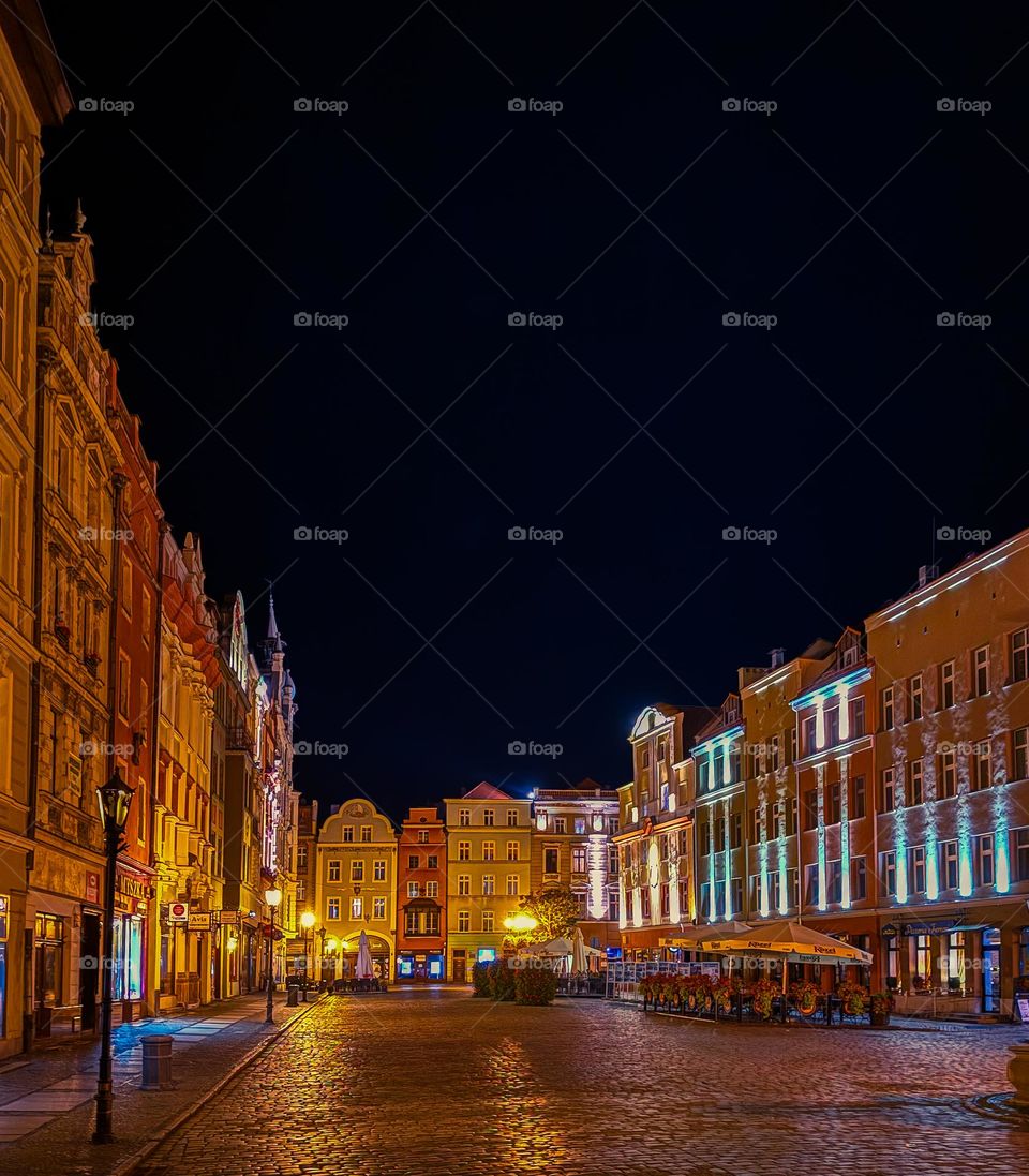 Night city square with lots of colorful small European houses and warm light, clear dark blue sky, no people