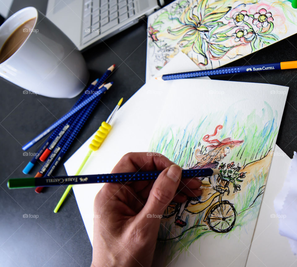 Hand holding Faber-Castell Aquarelle Colored watercolor pencils sketching girl on bicycle in countryside with sketchbook on table with laptop and coffee conceptual work life balance, creativity, hobby artist lifestyle photography 