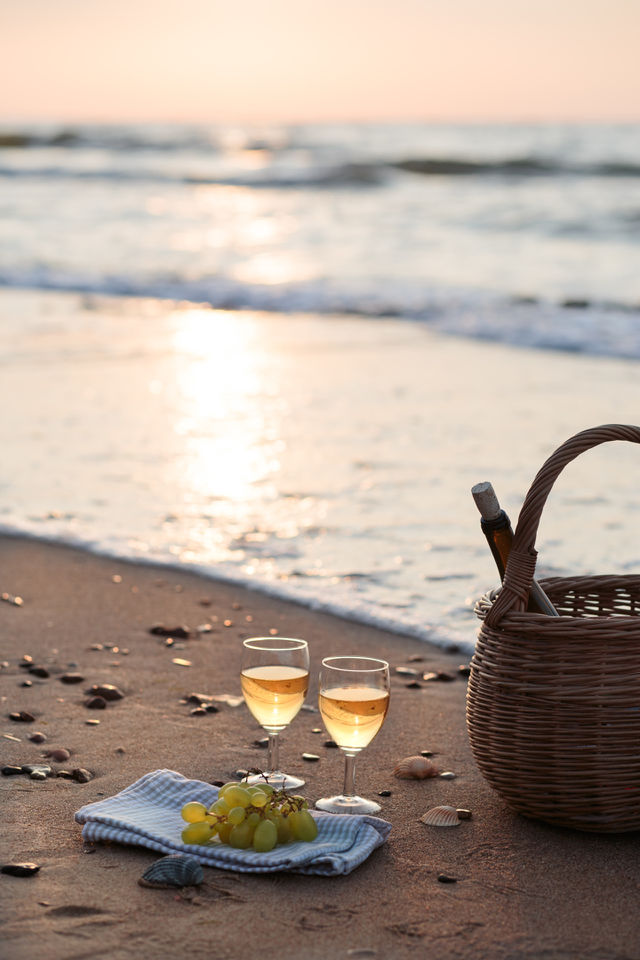 Two wine glasses with white wine standing on sand, on beach, beside grapes and wicker basket with bottle of wine. Sea waves in the background