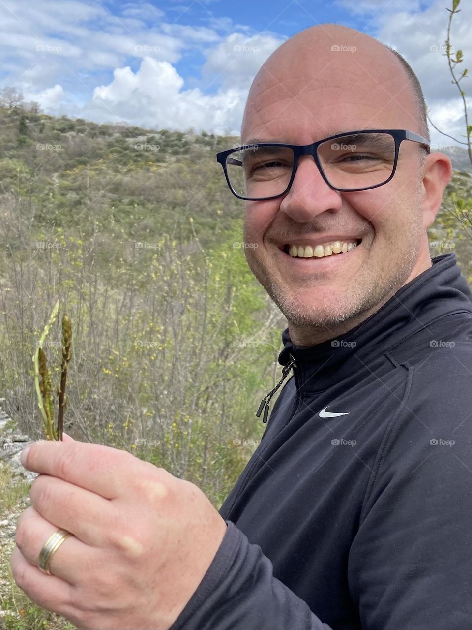 A bald headed man with glasses is smiling with sweet joy of success for foraging for wild asparagus.