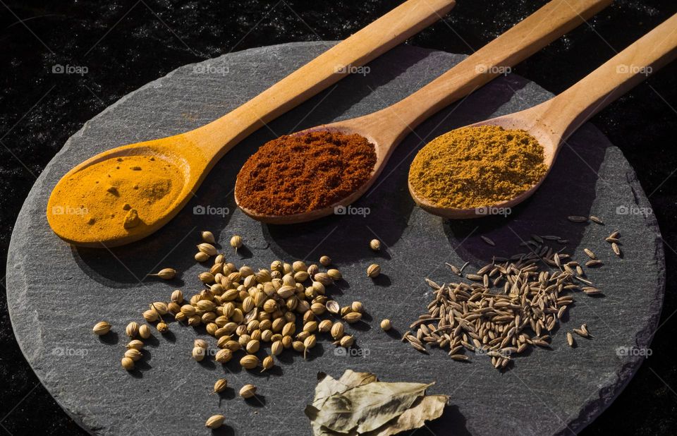 3 wooden spoons filled with spices in golden yellows, orange and red tones with seeds and leaves laid on grey slate