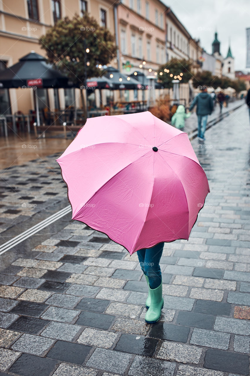 Child hiding behind big pink umbrella walking in a downtown on rainy gloomy autumn day