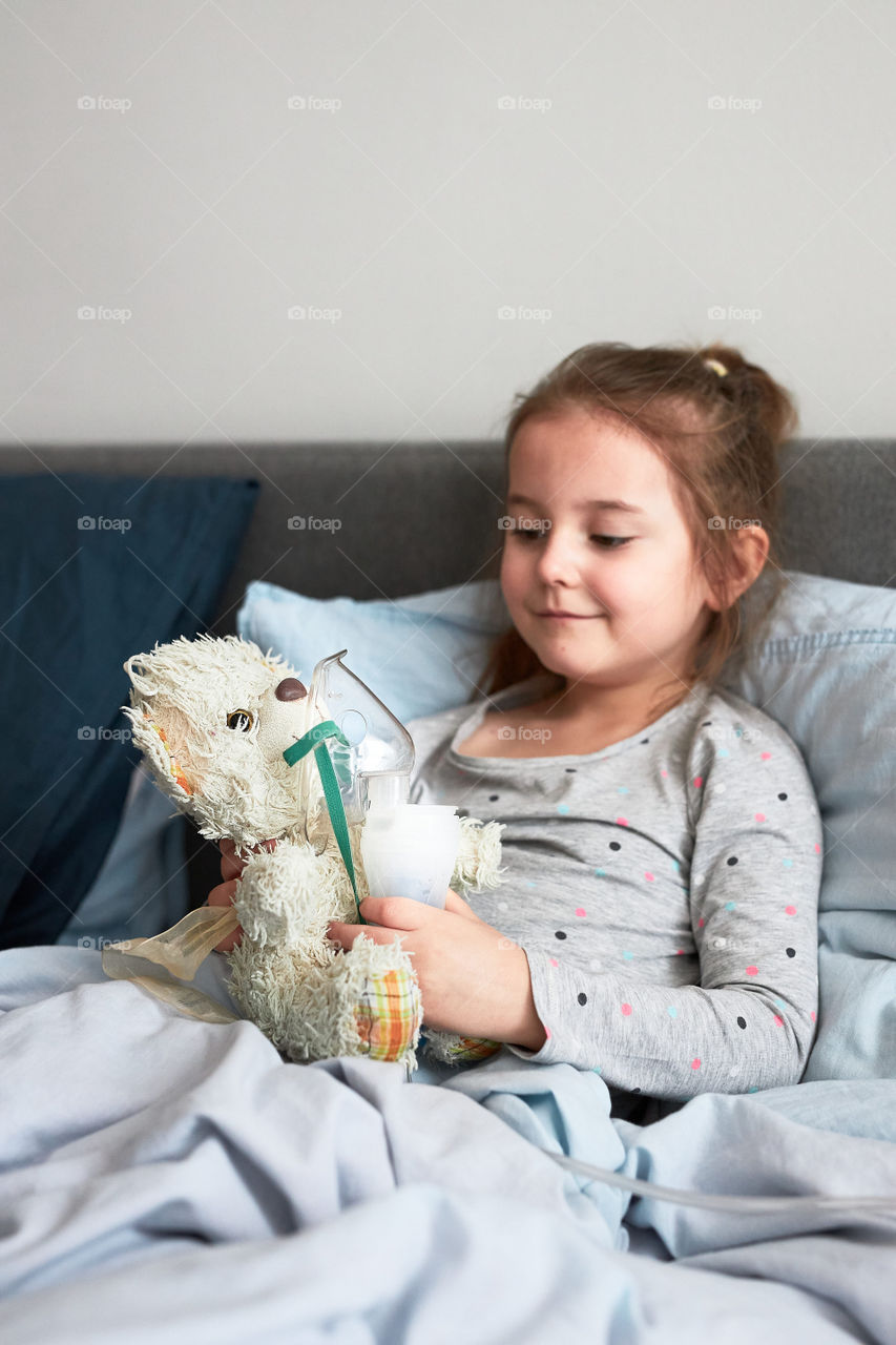 Sick child recovering in bed. Little girl playing by applying medical inhalation treatment with nebuliser to her teddy bear