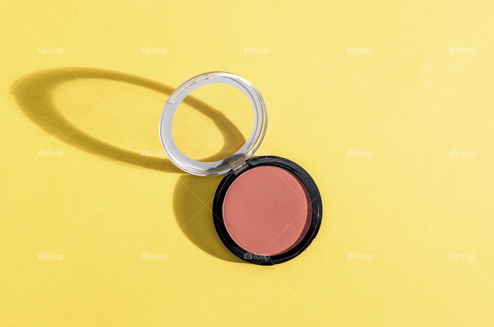 One round tonal dark pink face powder with an open transparent lid lies on a yellow background with hard shadows from the sun, close-up top view. The concept of female cosmetics, beauty.