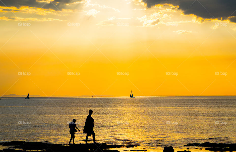 two people walking shoreline with yachts in distance silhouetted by the golden glow of the setting sun
