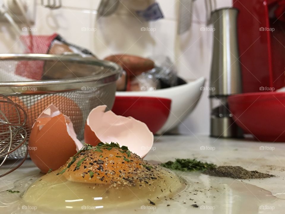 Cracked and seasoned raw egg on marble board, stainless steel colander filled with brown eggs in background, small piles of parsley and pepper