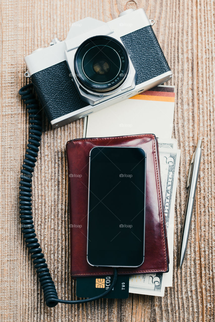 Smartphone with blank screen, camera, wallet, dollar banknotes, debit credit cards and notebook on wooden table. View from above. Portrait orientation