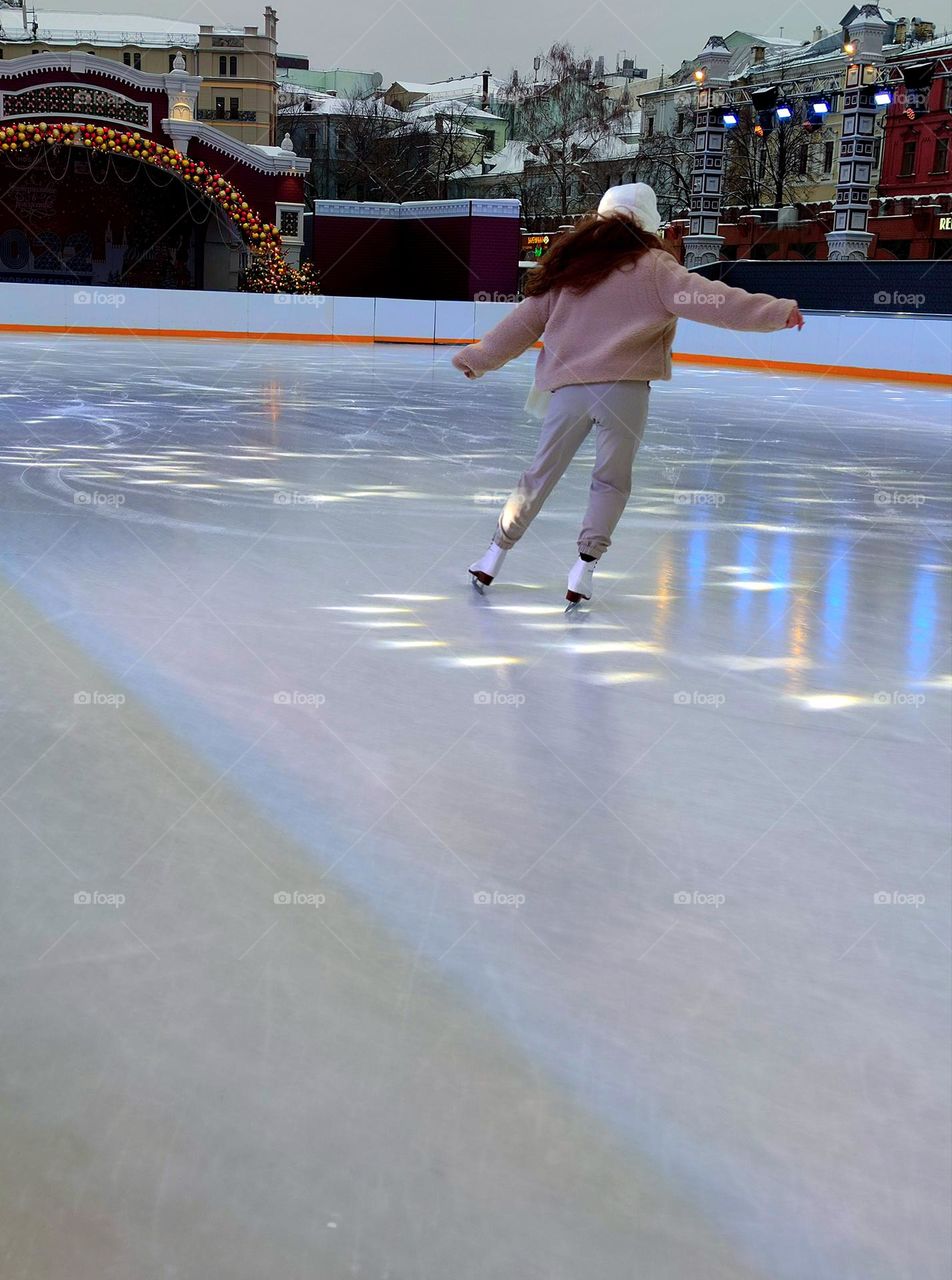 Winter.  A girl is riding a skating rink.  Multicolored light from searchlights falls on the ice.  Ice reflects nearby buildings.  Buildings in the snow around the ice rink