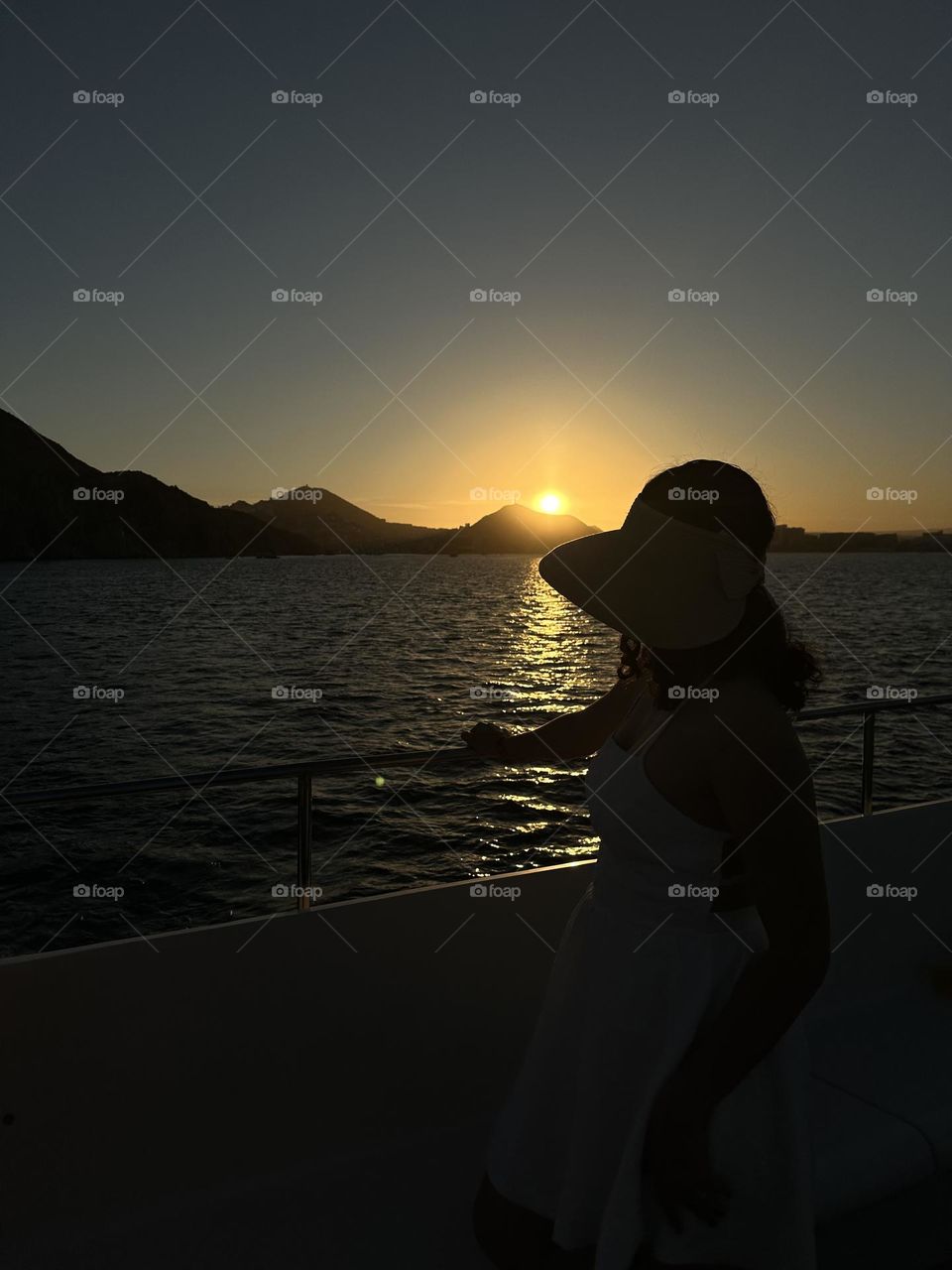 Woman wearing a hat looking at a beautiful sunset, while standing on a boat at the ocean.