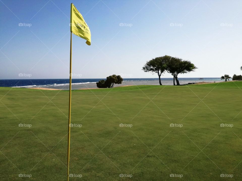 A beautiful view from behind the golf flag on the green to the beautiful blue sea and seashore.