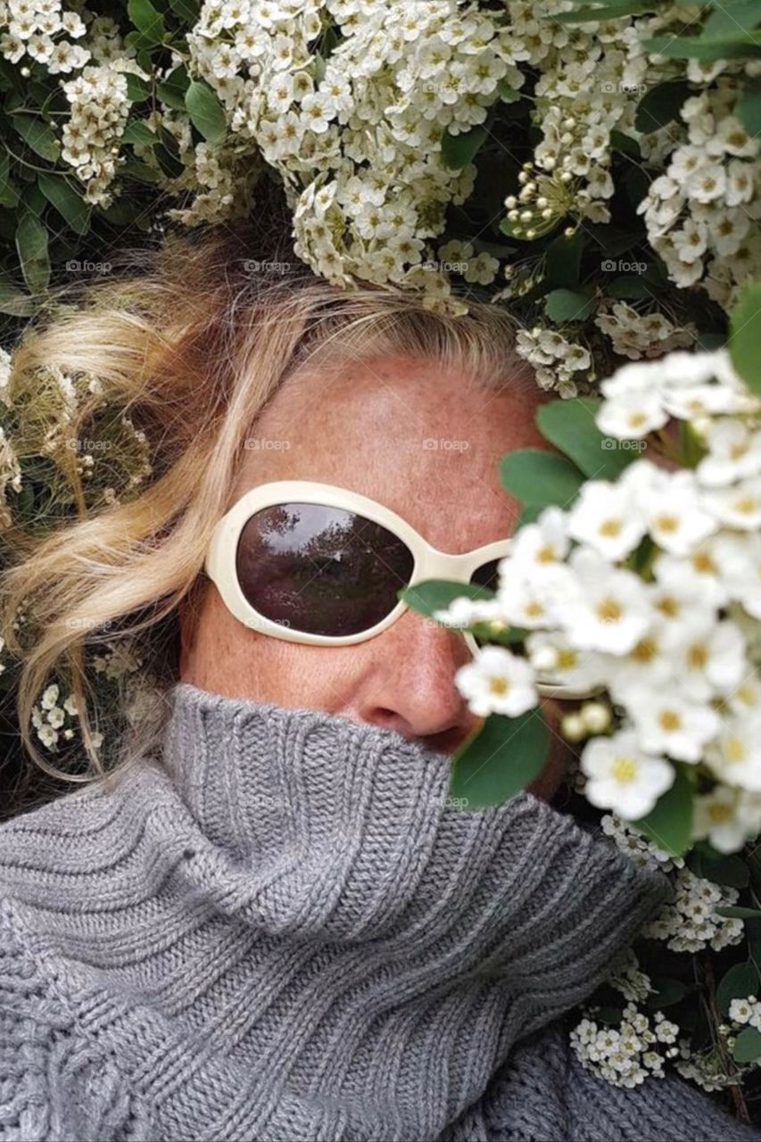 Face of a woman with freckles and white sunglasses and a thick turtleneck sweater between white flowers 