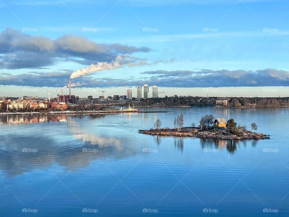 Tranquil scene with Helsinki coastline cityscape with skyscrapers, smoking pipe, little island with trees and small wooden cottage and clouds reflected in the smooth water surface of Baltic Sea 