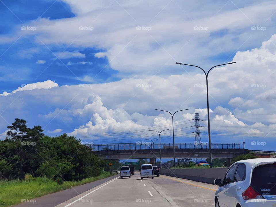 Driving on the toll road, the weather is sunny, the sky refract blue color, the clouds sppear pure white, shady green leaf trees as barrier to surrounding environment