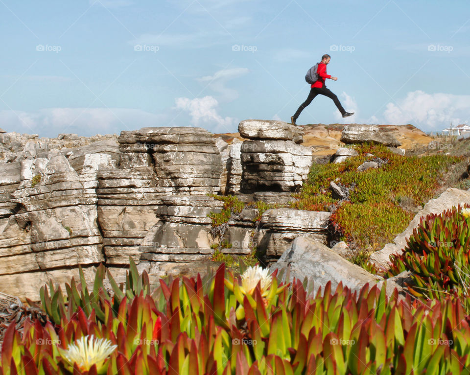 A man in a red shirt and wearing a backpack leaps over a gap between some rocks on a clear, bright morning, the red tips of plants in the foreground echo colour of the man’s shirt