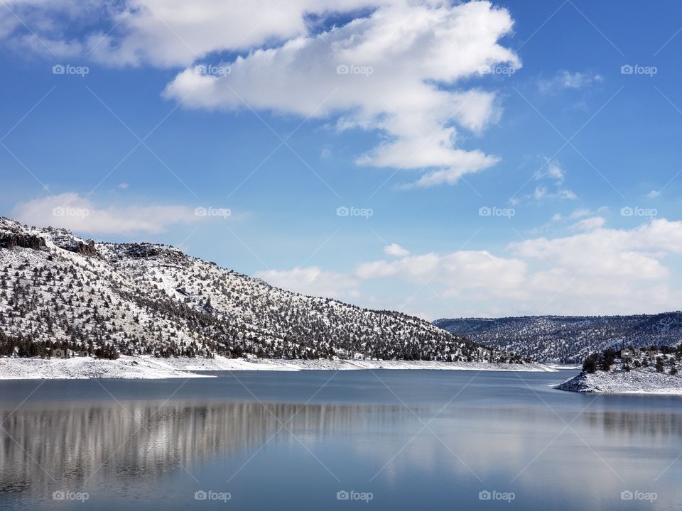 Snow covered hills, trees, clouds, and a bright blue sky reflect in the partially iced over Prineville Reservoir on a sunny winter day in Central Oregon.