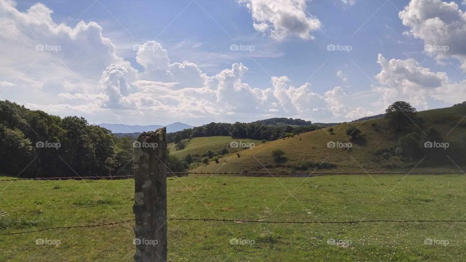 Pasture with orange and green hills and trees behind, a wire fence and wooden posts in front, blue mountains in the distance, and bright blue sky with billowing white clouds above.