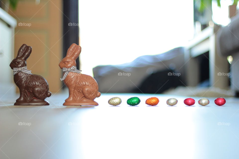 A portrait of two chocolate easter bunnies followed by a row of chocolate easter eggs. the eggs are wrapped in different colors of tin foil.