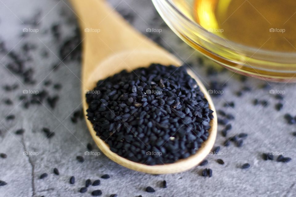 Closeup of black seed in a bamboo spoon next to a small glass bowl of oil on a stone surface