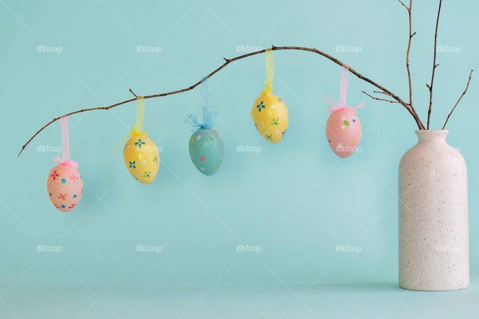 Decorative Easter eggs hanging on dry branch placed in white ceramic vase with two blue eggs near against blue background