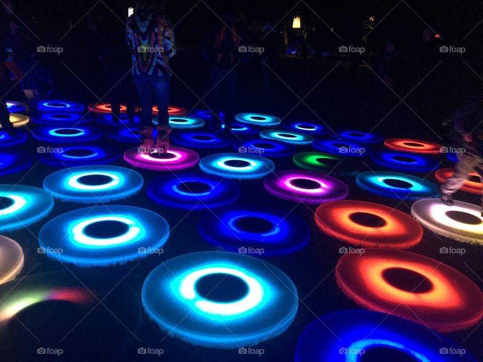 Glowing blue circles in a pattern on the ground. There are a few in other colors scattered among them. A few people stand in the background. 