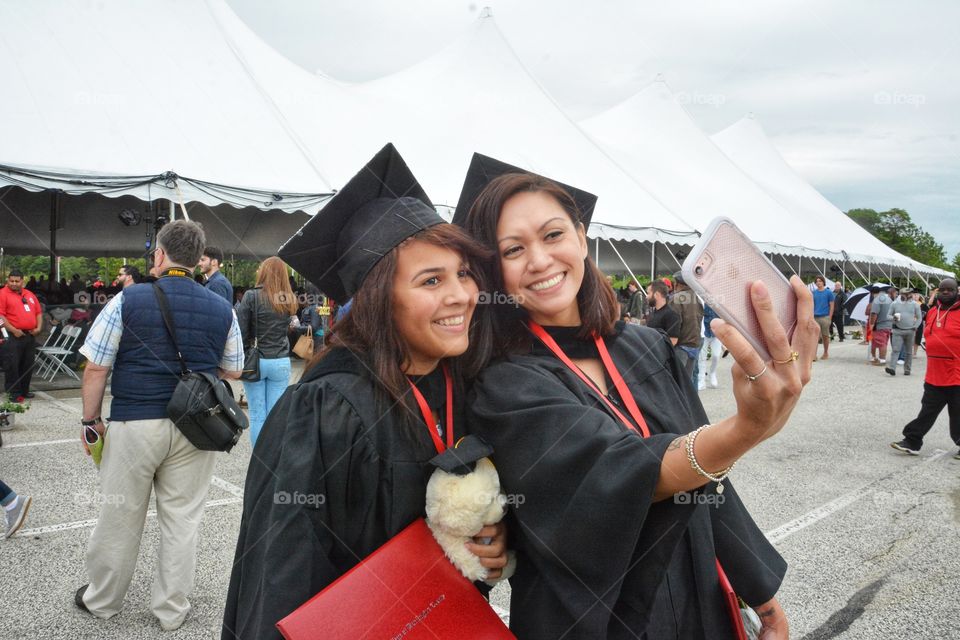 Two young women are seen wearing a cap and grown during graduation, taking a selfie photo with a cell phone.