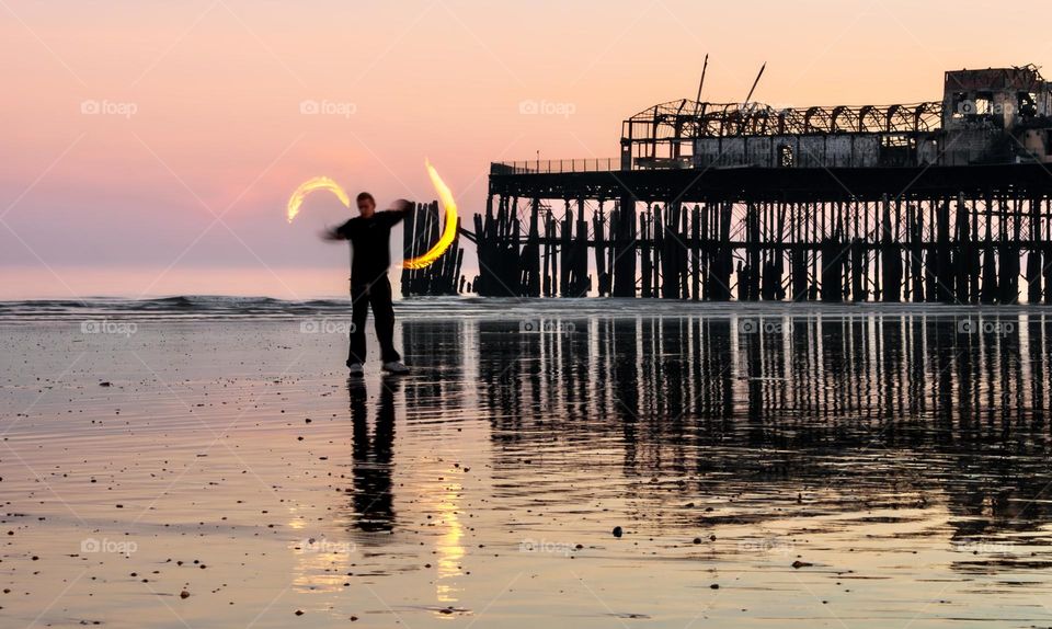 A man spins fires poi on a wet, sandy beach at sunset in front of the remains of an old pier.
