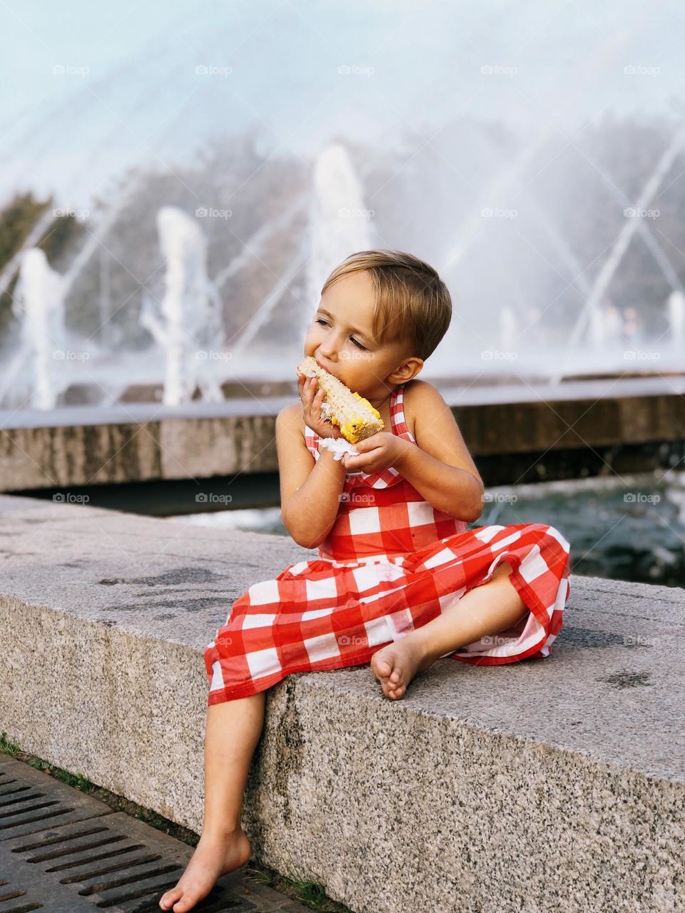 Cute little girl with short blonde hair wearing red dress sitting near fountains and eating corn, portrait of child 
