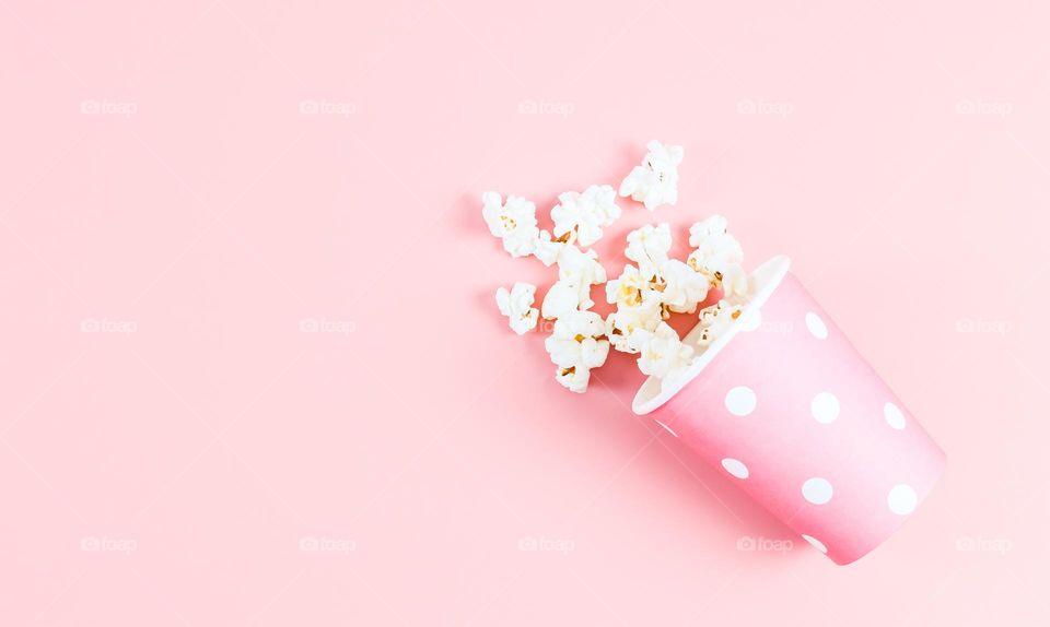 A single pink polka dot paper glass with popcorn popping out of it is on the right against a soft pink background with copy space on the left, flat lay closeup. Cinema concept, disposable tableware with polka dots.