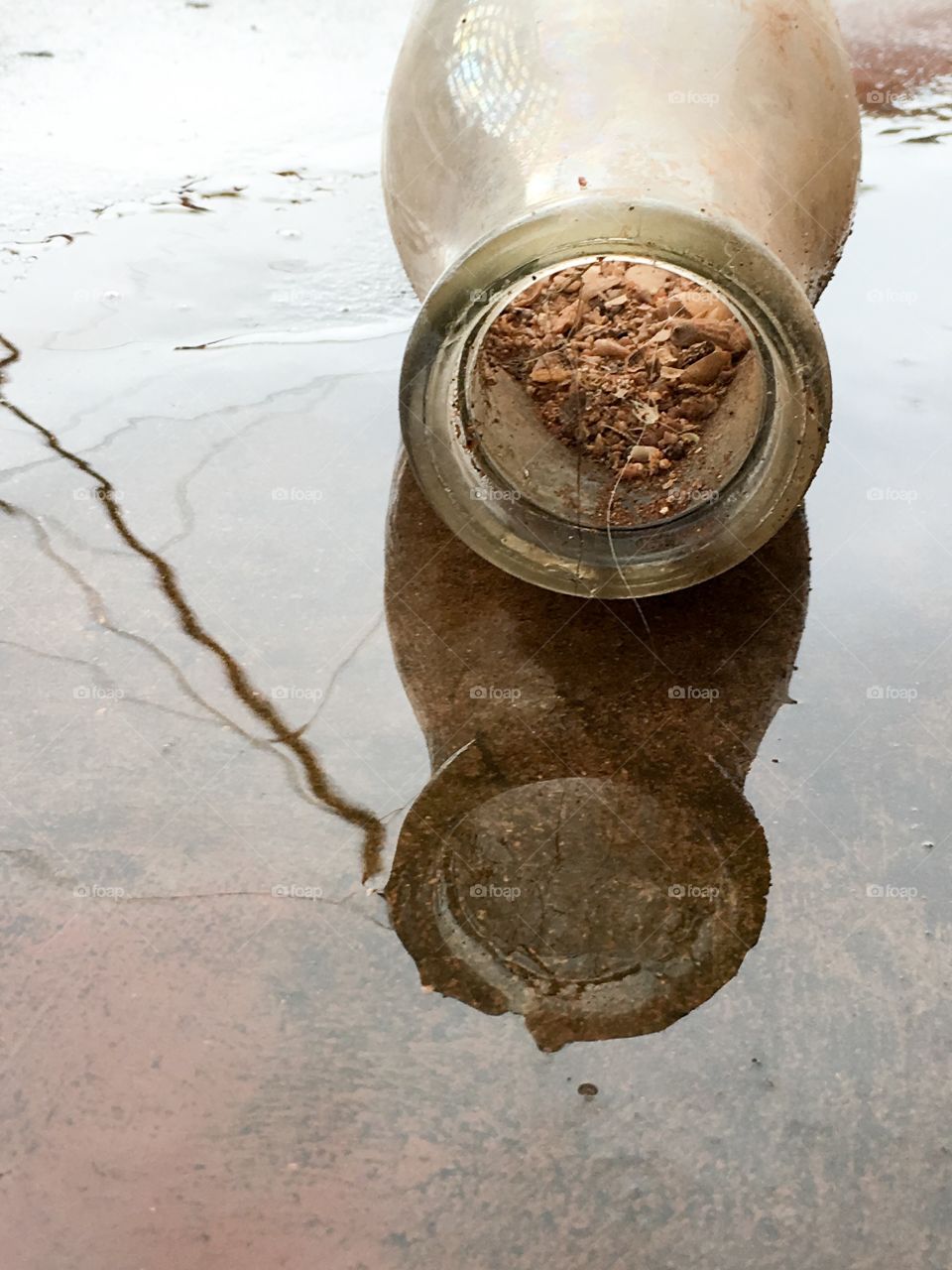 Antique milk bottle filled with dirt sand and seashells as found when dug up, laying on its side in the rain and rainwater and its reflection 