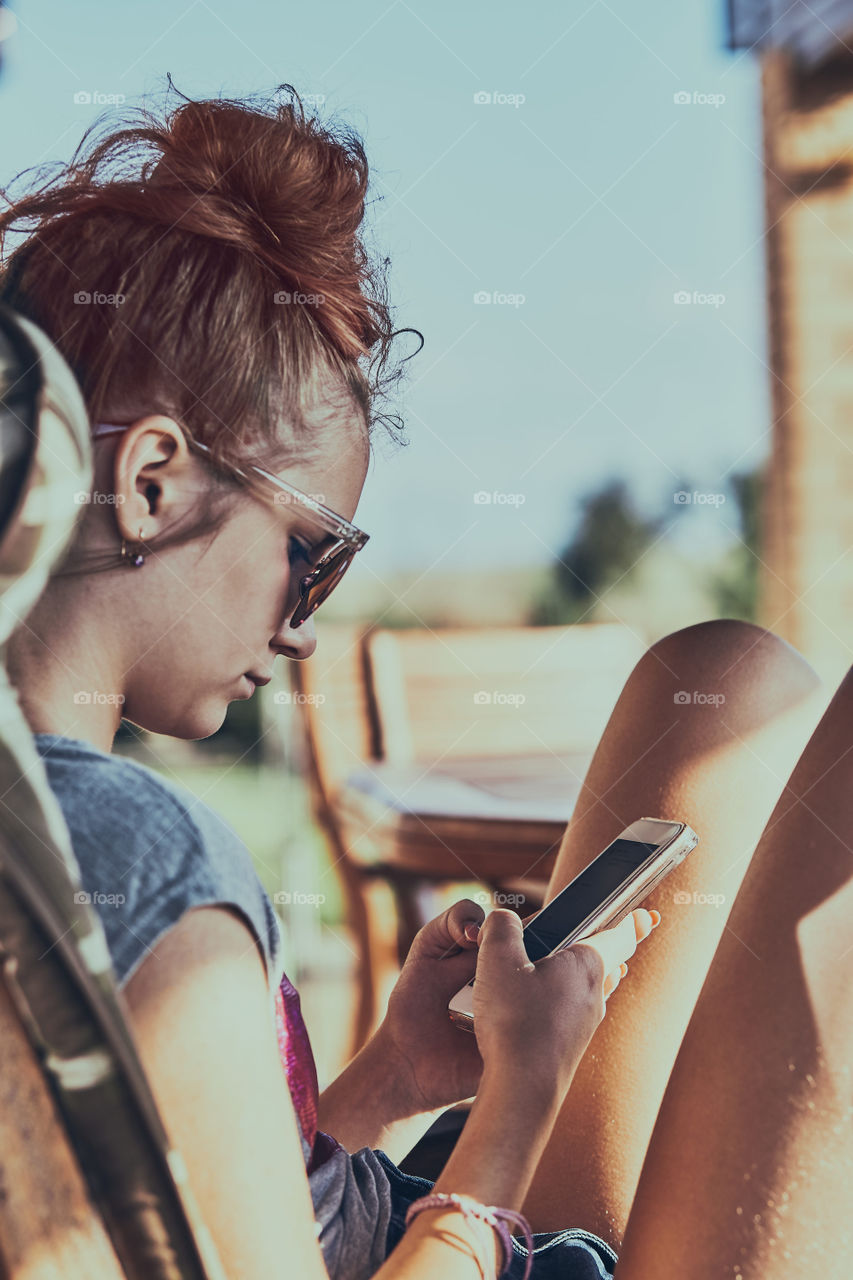 Young woman chatting on a  mobile phone sitting in a chair outdoors on patio wearing a sunglasses. Real people, authentic situations