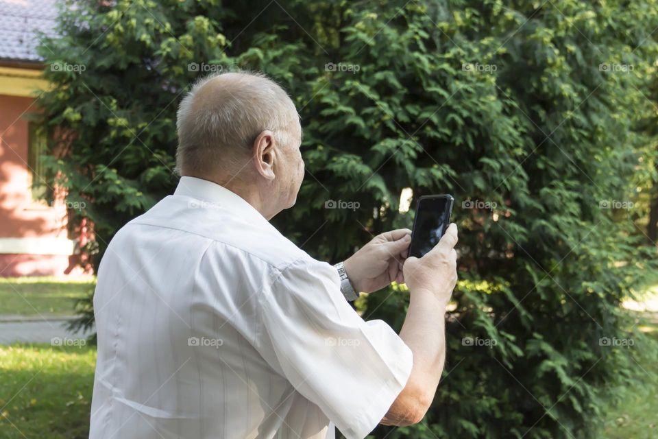An elderly man walks alone in the park in the summer. A modern pensioner, businessman in a white shirt and trousers takes pictures with a camera in a mobile phone.