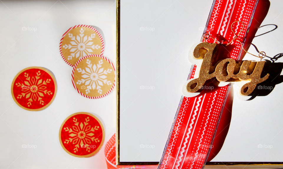 A white and gold rimmed box waits to be wrapped up with a red Christmas ribbon and a joy decoration