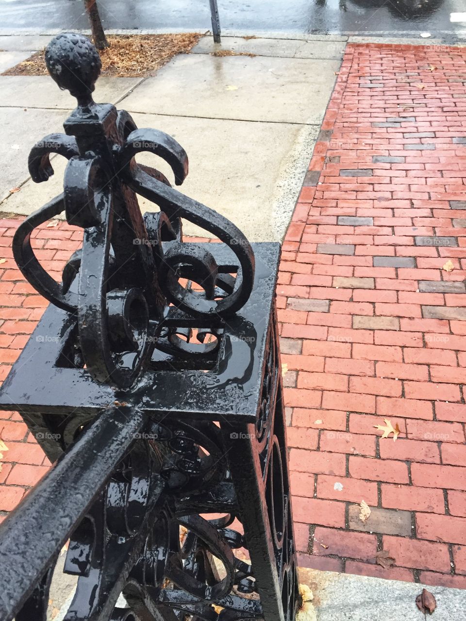 Decorative iron work tops the post of a stair railing next to a fence. The wrought iron peace is painted black. It is covered with water because it is raining out.