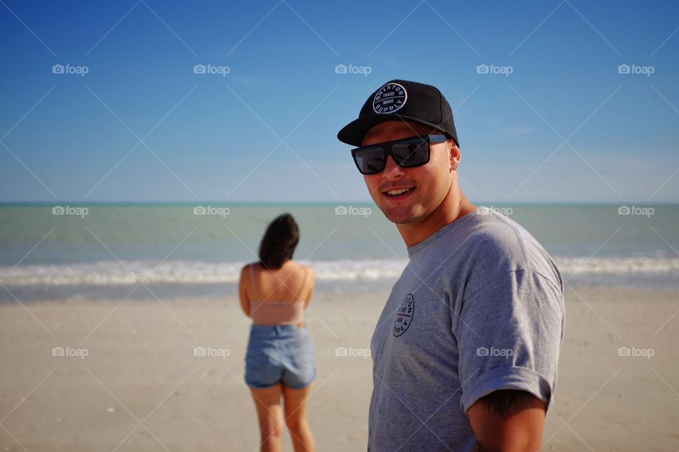 Man smiling at camera with woman in background gazing over the ocean