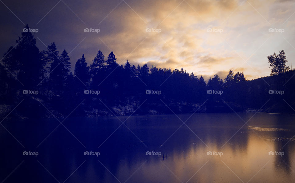 Winter landscape. Frozen lake reflects the evergreen trees surrounding it as well as the clouds. Colourful sky above. 