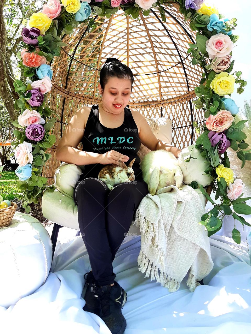 Young lady lovingly petting bunnies, seated, surrounded with flowers of pastels colors representing Easter.