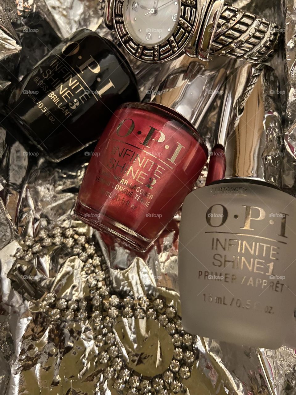 Opi Infinite Shine Nail Lacquer in shade “15 Minutes of Flame” along with the primer & top coat nestled in the middle of a silver iridescent background between a bracelet shaped in the infinity symbol & a silver watch.
