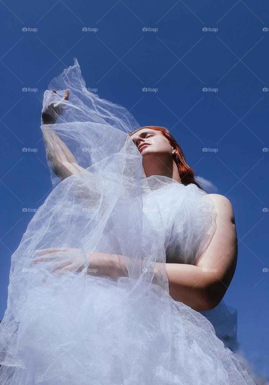 a white-skinned girl with red hair in a white chiffon in the form of a white cloud against a blue sky