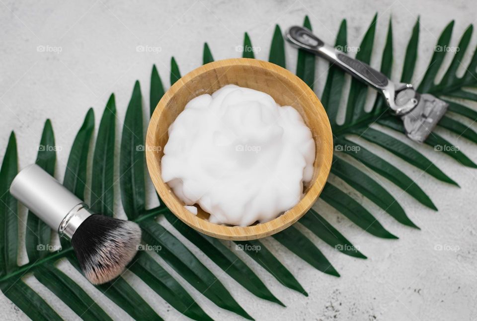 One wooden bowl of shaving foam, shaving brush and razor lie on a palm tree diagonally on a light cement background, flat lay. Concept barbershop man, shaving man, beard care.