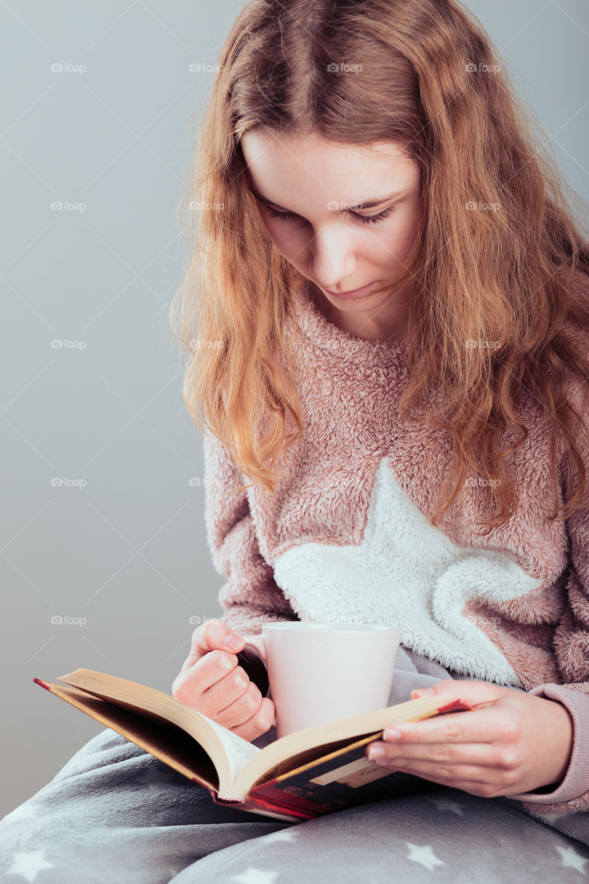 Girl enjoying the reading a book and drinking coffee at home. Young woman sitting on a chair, wrapped in blanket, holding book, relaxing at home. Portrait orientation. View from above